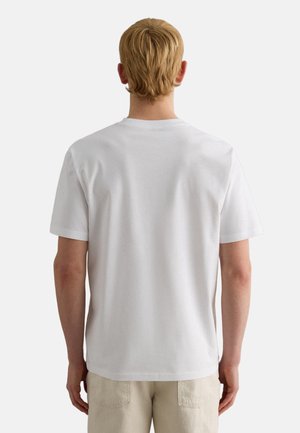 Blonde persoon staand met de rug naar de kijker, gekleed in een effen witte T-shirt met korte mouwen en lichtbeige broek tegen een effen achtergrond.