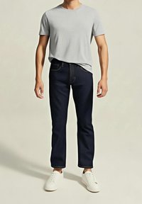 Homme portant un t-shirt gris clair à manches courtes, un jean bleu foncé et des baskets blanches, debout devant un fond clair uni.