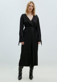Schwarzes Wickelkleid mit langen Ärmeln, tailliertem Gürtel, strukturiertem Stoff und Fransen Details an den Ärmeln. Tragen mit kniehohen schwarzen Stiefeln.