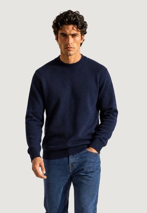 Uomo con capelli scuri e ricci che indossa un maglione blu navy e jeans blu, in piedi con una mano in tasca davanti a uno sfondo uniforme.