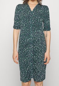 Femme portant une robe verte à fleurs, mi-longue, avec un devant froncé et des manches trois-quarts, se tenant devant un fond uni.