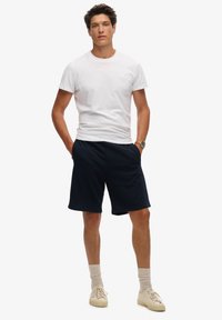 T-shirt bianco in cotone e pantaloni corti blu navy, con una vita elastica. Sneakers color crema e calze grigie chiare completano l'outfit.