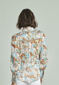 Blusa a maniche lunghe in tessuto leggero con un motivo astratto blu e marrone. Presenta pieghe e dettagli in catena sui polsini.