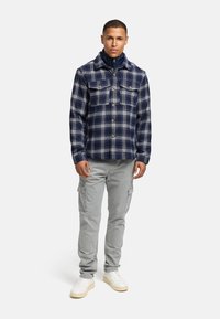 Chemise en flanelle bleu marine avec un motif à carreaux blancs, deux poches poitrine, boutons en métal ; associée à un pantalon cargo gris clair et des baskets blanches.