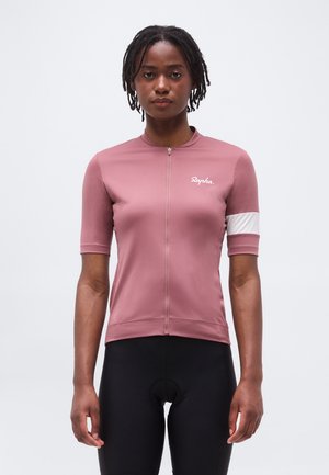 Γυναίκα που φορά ροζ ποδηλατική φανέλα με κοντά μανίκια, εμπρόσθιο φερμουάρ και λευκή ρίγα στο μανίκι, συνδυασμένη με μαύρη ποδηλατική βερμούδα.