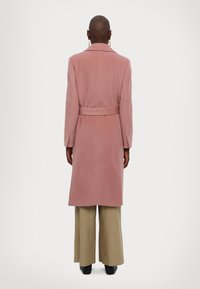 Lång rosa kappa med bälte, med en notchad krage och skräddarsydda ärmar, som bärs över vida ljusa byxor och svarta stövlar.