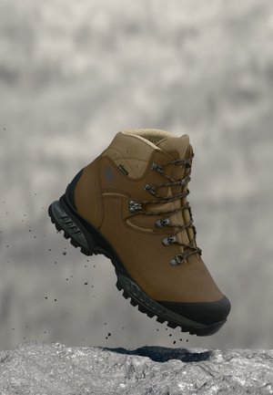 Brauner Wanderschuh mit schwarzer Sohle und verstärkter Spitze, schwebt in der Luft über felsigem Untergrund mit fallenden kleinen Steinen.