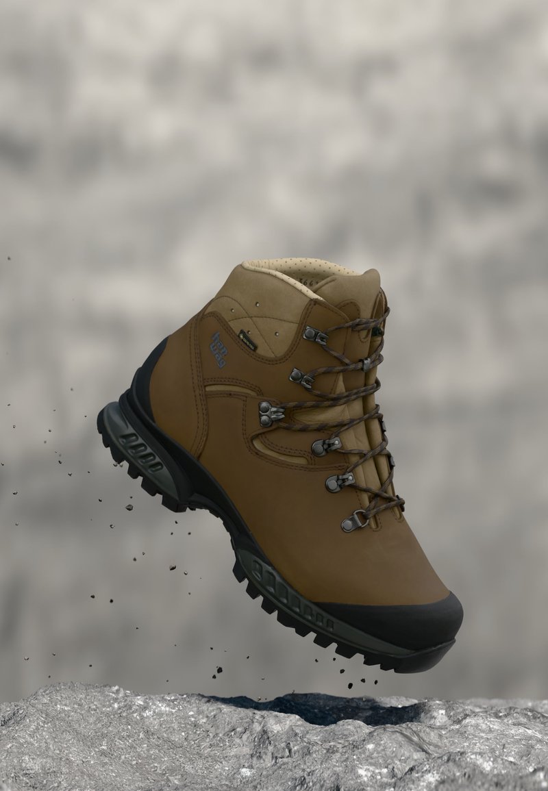 Καφέ ορειβατικό μπότες με μαύρη σόλα και ενισχυμένη μύτη, αιωρείται στον αέρα πάνω από βραχώδες έδαφος με πέτρες που πέφτουν.