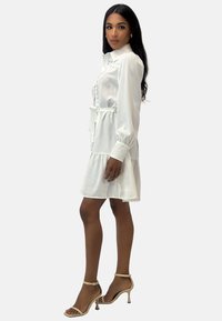 Robe en satin blanc à manches longues, boutons sur le devant et taille ceinturée. Elle présente une jupe à étages et une broderie subtile le long du col.