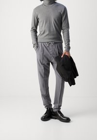 Grauer Rollkragenpullover, gestreifte graue Hose mit Kordelzug und schwarz Schnürschuhe. Hält ein schwarzes Blazer.