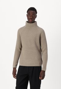 Beige gestrickter Rollkragenpullover mit gerippter Textur, ausgestattet mit Raglanärmeln und einem figurnahen Design. Getragen mit schwarzen Hosen.