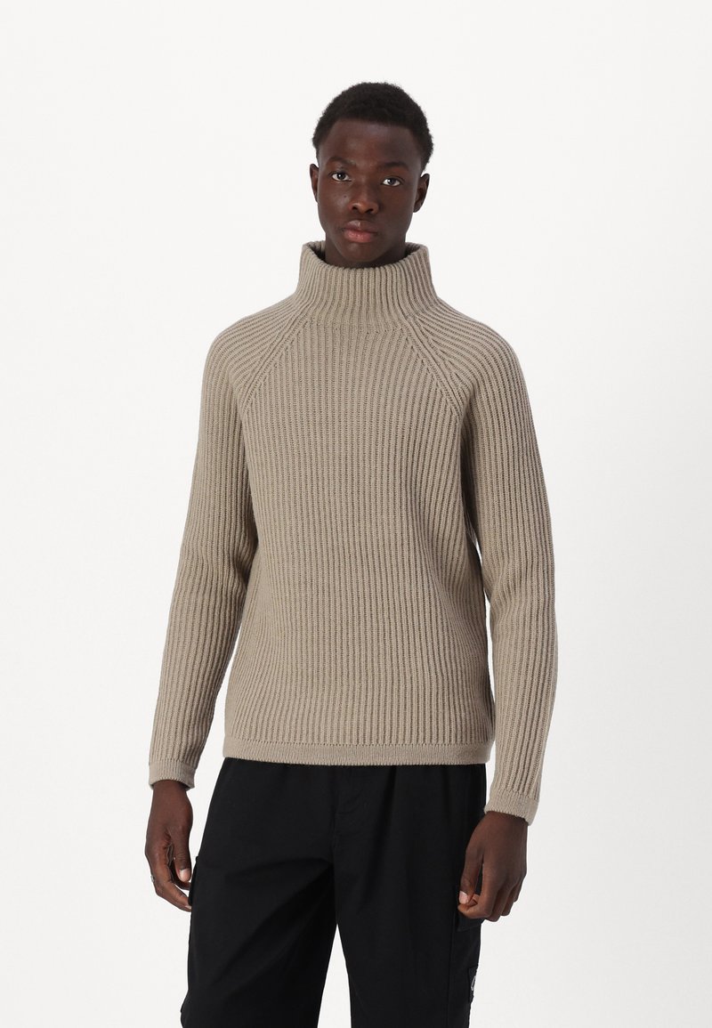 Beige gestrickter Rollkragenpullover mit gerippter Textur, ausgestattet mit Raglanärmeln und einem figurnahen Design. Getragen mit schwarzen Hosen.