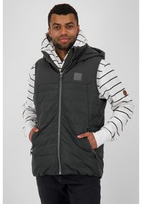 Gevoerde zwarte vest met capuchon, voorzien van een ritssluiting aan de voorkant, twee zijzakken en een vierkant patch. Daaronder een wit gestreepte long-sleeve hoodie.
