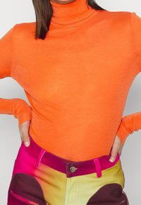 Person klädd i en ljusorange polotröja och färgglada byxor med rosa, gula och mörklila lappar, med händerna på höfterna.