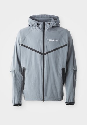 Light gray windbreaker jacket with a hood. Features black accents, a front zipper, and two side pockets. "BARCA" and Nike logo printed on front.