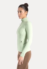 Chaqueta verde claro con cremallera, cuello alto, mangas largas y dos bolsillos laterales con cremallera, fabricada con una tela lisa y elástica.