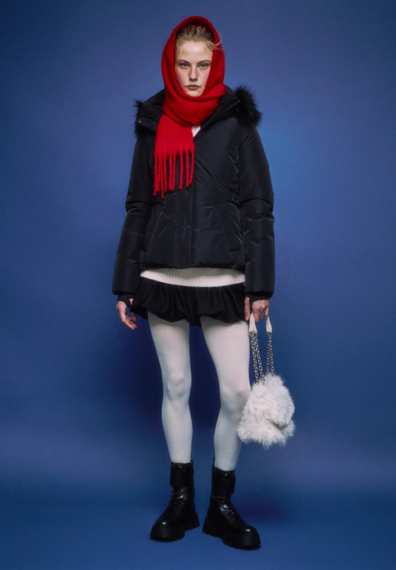 Črna podložena jakna s krznom na robu, rdeč šal, beli pulover, črno krilo z volančki, bele nogavice, črni škornji, drži puhasto belo torbico.