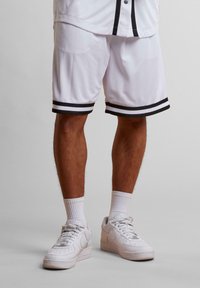 Shorts de basketball blancs avec des rayures noires au niveau de l'ourlet, fabriqués en tissu léger. Associés à des baskets blanches et des chaussettes blanches côtelées.