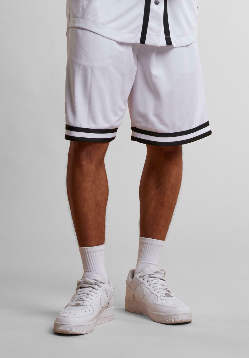 Shorts de basketball blancs avec des rayures noires au niveau de l'ourlet, fabriqués en tissu léger. Associés à des baskets blanches et des chaussettes blanches côtelées.
