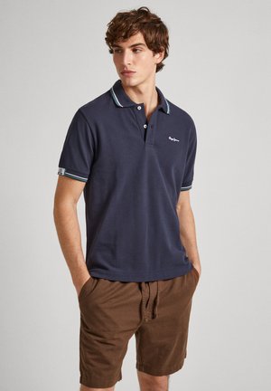 Camisa polo de color azul marino con mangas cortas, cuello en contraste y logo de la marca. Combinada con pantalones cortos color marrón claro y manos en los bolsillos.