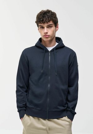 Giovane uomo con capelli ricci che indossa una felpa con zip blu navy e pantaloni beige, in piedi contro uno sfondo chiaro e uniforme.