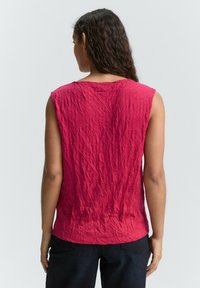 Femme aux longs cheveux bouclés portant un haut rouge sans manches froissé et un pantalon sombre, vue de dos sur un fond uni.