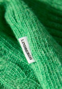 Grön stickad tyg med en mjuk textur. En vit etikett med "LINDBERGH" sydd i tyget. Närbild på materialdetaljer.