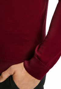 Burgunderfarbener Strickpullover mit glatter Textur, gerippten Bündchen und Saum. Zeigt eine Hand in einer schwarzen Tasche, die ein geschichtetes Design zur Schau stellt.