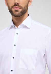 Camicia bianca con colletto a punta, dettagli blu sui bottoni e una tasca singola sul lato sinistro del petto. Texture del tessuto liscia.