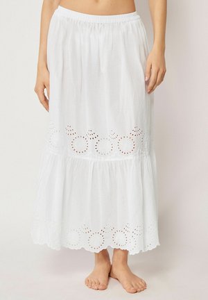 Frau trägt einen weißen knöchellangen Rock mit elastischem Bund und bestickten Lochblumenmustern am Saum. Barfuß.
