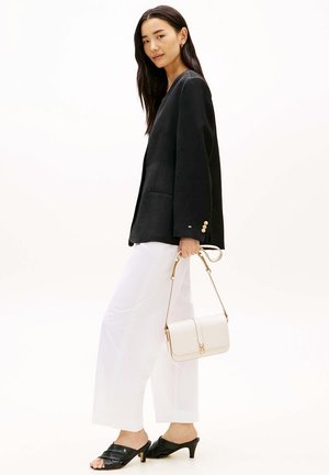 Femme en blazer noir, pantalon blanc à jambes larges, sandales noires à talons, tenant un sac à main blanc sur fond blanc.