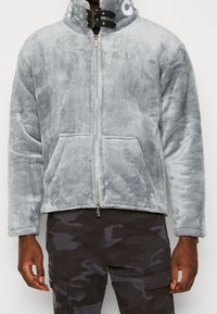 Szara kurtka polarowa z wysokim kołnierzem, z przodu na zamek błyskawiczny i dwie przednie kieszenie; posiada czarne detale i subtelne logo na kołnierzu.