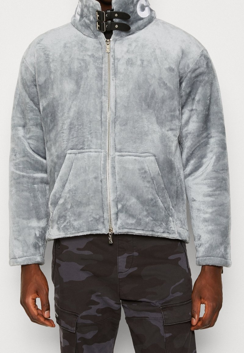 Szara kurtka polarowa z wysokim kołnierzem, z przodu na zamek błyskawiczny i dwie przednie kieszenie; posiada czarne detale i subtelne logo na kołnierzu.