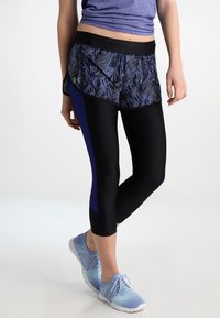 Kvinnors träningsoutfit med mönstrade shorts i svart och blått över svarta leggings, samt blå sneakers. Materialet verkar elastiskt och andningsbart.