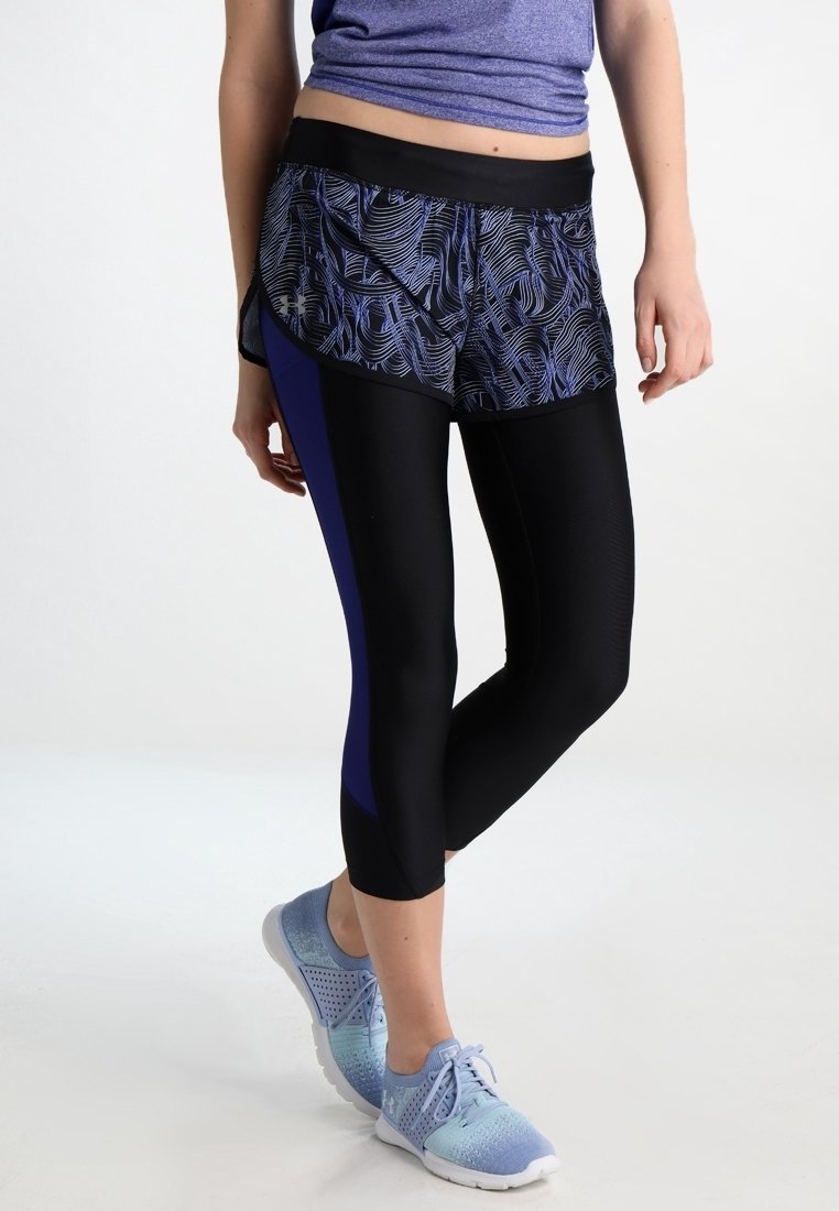 Kvinnors träningsoutfit med mönstrade shorts i svart och blått över svarta leggings, samt blå sneakers. Materialet verkar elastiskt och andningsbart.