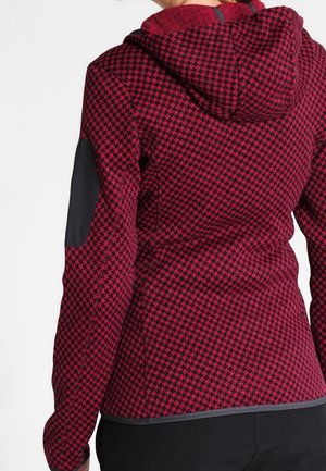 Vue de dos d'une personne portant un sweat à capuche à motif pied-de-poule rouge et noir avec des coudières noires et un pantalon noir.