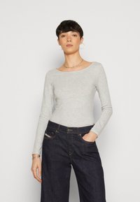 Helles graues Oberteil mit langen Ärmeln aus weichem Stoff, das einen weiten Ausschnitt hat. Kombiniert mit dunklen Denim-Hosen im Weitschnitt mit Fronttaschen.