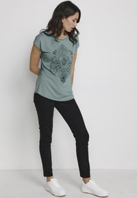 Turquoise t-shirt met zwart paisleyprint, korte mouwen, gecombineerd met zwarte skinny jeans en witte sneakers. Glad textuur.