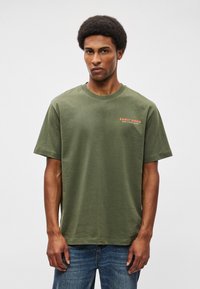T-shirt de algodão verde-oliva com um corte descontraído, gola redonda e texto "EARLY BIRDS" em laranja no lado esquerdo do peito. Tecido ligeiramente texturizado.