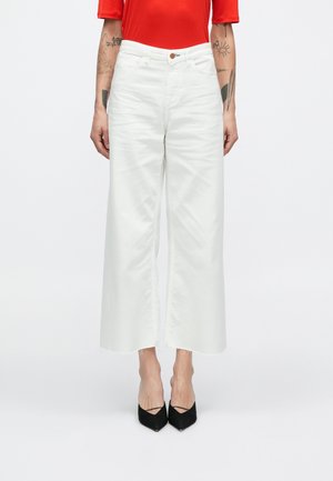 Hvite vide bukser laget av denim, med høy midje, frynsete kant og en brun knapp på linningen. Kombinert med svarte sko.