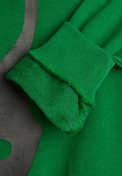 Πράσινο πλεκτό μανίκι με διπλωμένο μανσέτα πάνω σε φόντο υφασμάτινης πλέξης με υφή που διαθέτει γκρι καμπυλωτό σχέδιο.