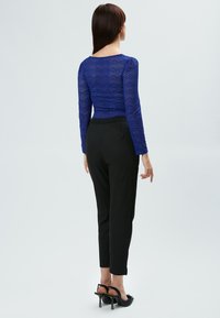 Blauw kanten langgledige top gecombineerd met zwarte strakke broek en zwarte hoge hakken, gezien van achteren.