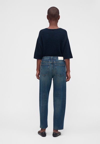 Personne aux cheveux courts portant un pull bleu marine à manches courtes, un jean droit bleu et des chaussures noires sans lacets, debout de dos.