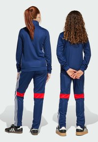 Twee kinderen in marineblauwe trainingspakken met rode accenten staan rug aan rug, weg van de camera. De een heeft lang, steil haar, de ander heeft krullend haar.