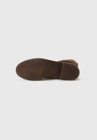 Bottines en suede marron avec une semelle en caoutchouc texturée, dotées d'un bout rond et de détails de piqûre sur les côtés. La semelle extérieure présente une surface de grip à motifs.