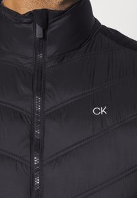 Chaqueta aislante negra con patrón en forma de chevron, cuello alto y cremallera frontal. Presenta un pequeño logotipo blanco "CK" en el lado izquierdo.