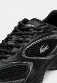 Chaussure de sport noire avec des empiècements en mesh, des lacets épais et un logo crocodile argenté sur le côté, soulignant son design sportif.