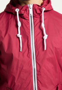 Chaqueta ligera roja hecha de material brillante con cremallera completa, cordones blancos y capucha. Presenta un diseño simple y sin patrones visibles.