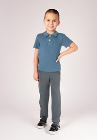 Niebieska koszulka polo z kołnierzykiem i guzikiem, w połączeniu z szarymi spodniami. Czarne trampki z białymi podeszwami. Dziecko stoi, trzymając jedną rękę na biodrze.