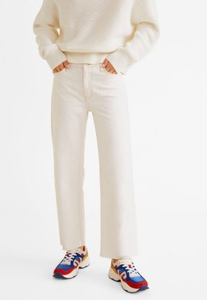 Person trägt cremefarbene Jeans mit weitem Bein, einen cremefarbenen Strickpullover und bunte klobige Sneaker mit blauen, roten und senffarbenen Akzenten.